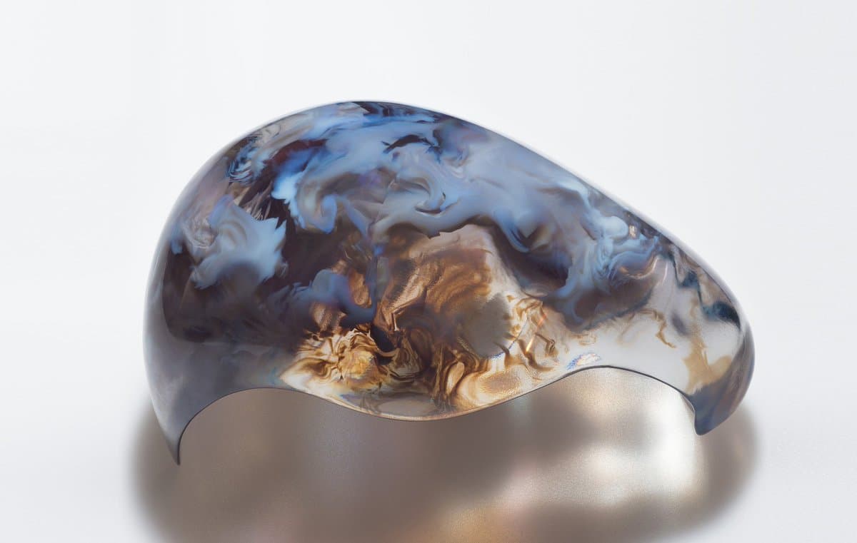 A studio photograph of a glossy, sculptural object with a smooth, curved form and a marbled surface pattern in shades of blue, white, and brown, set against a plain white background.