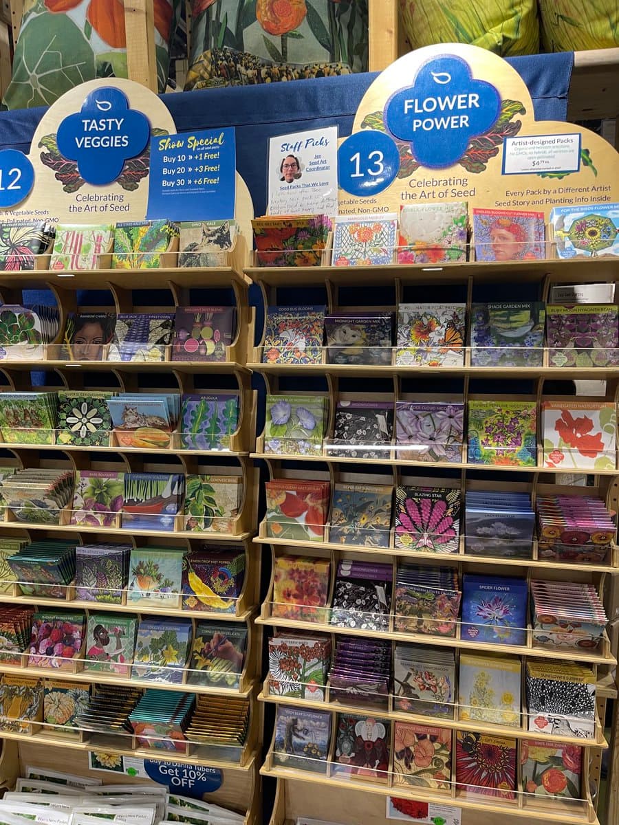 A retail display of artist-designed seed packets organized on wooden shelves, featuring promotional signs for 'Tasty Veggies' and 'Flower Power' sections. The colorful packaging showcases various floral and vegetable illustrations, with a 'Staff Picks' sign highlighting a specific selection.