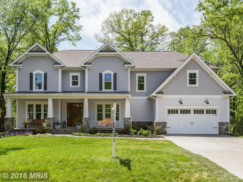 A real estate photograph of a large, two-story house with light gray siding, white trim, and a white garage door, set on a green lawn with trees in the background.