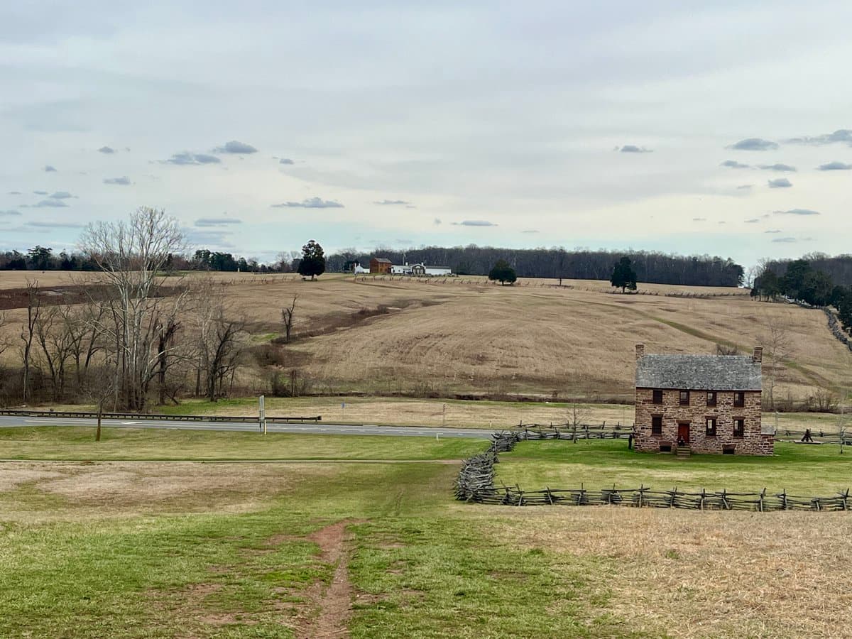 A wide landscape photograph showing a historic stone house with a slate roof and two chimneys, surrounded by a split-rail fence and green grass. In the background, there are rolling hills with dry, brown grass, a few scattered trees, and distant farm buildings under an overcast sky.