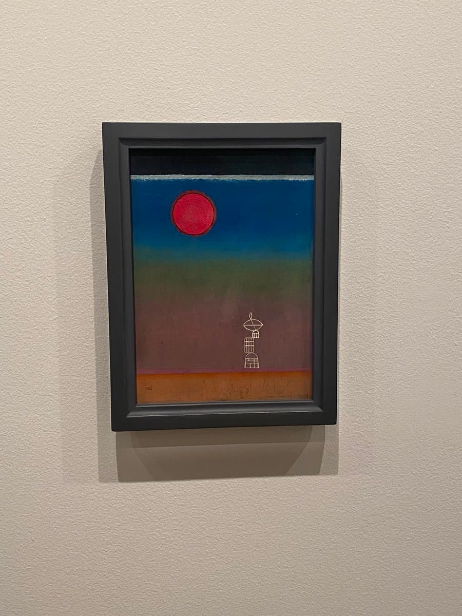 A framed painting hanging on a white wall, depicting a stylized landscape with a large red sun in a blue sky and a white line-drawing of a satellite dish on a brown ground.