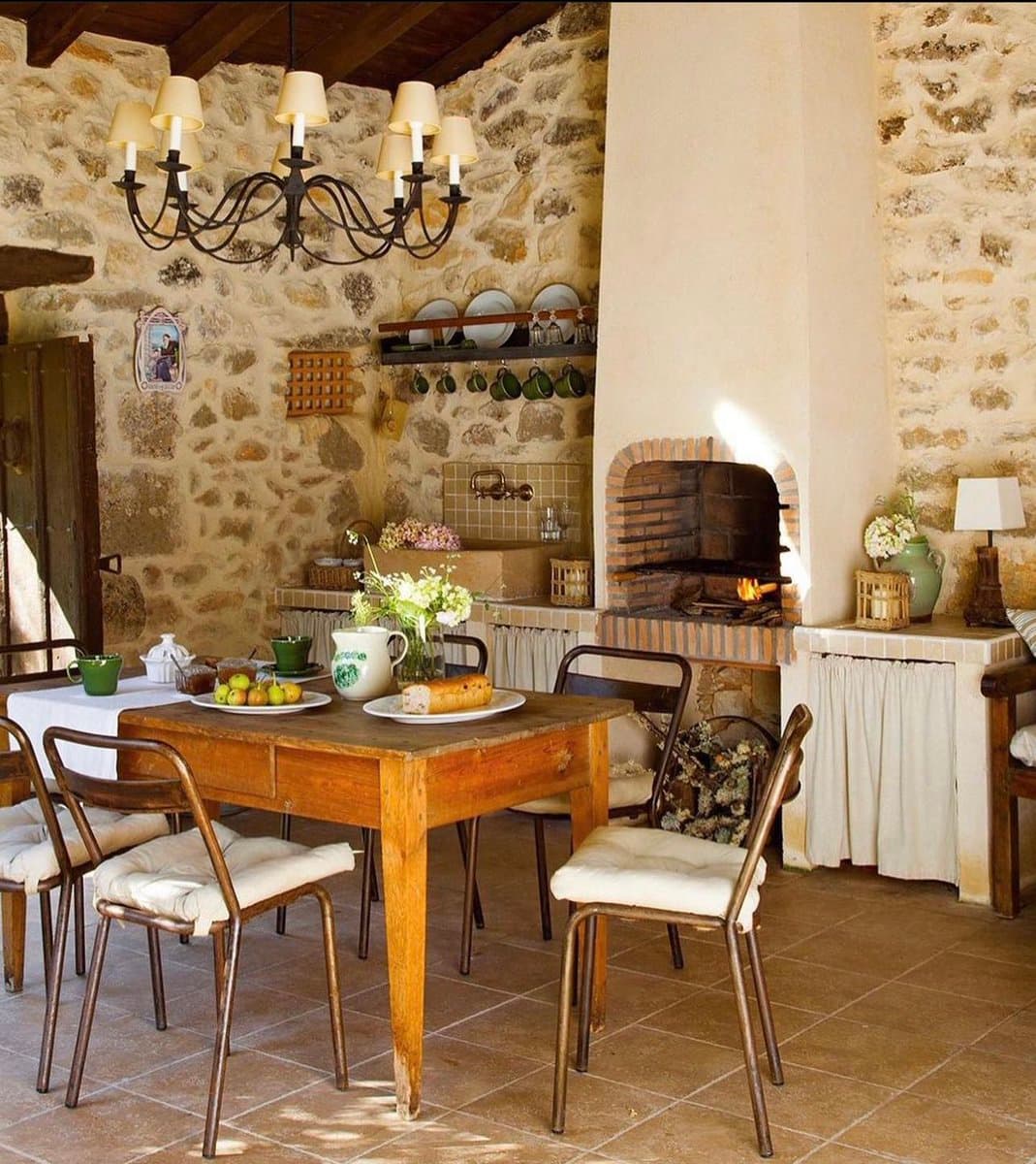A rustic dining room featuring a wooden table set with bread, fruit, and flowers, surrounded by metal chairs with white cushions. The room has exposed stone walls, a large brick fireplace with a fire burning, and a wrought-iron chandelier hanging from the ceiling.
