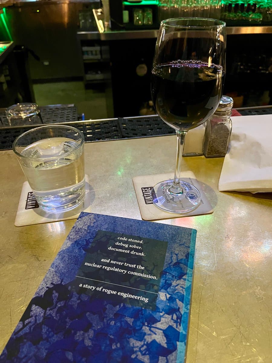 A photo of a book titled 'Midnight Simulacra' resting on a bar counter next to a glass of red wine and a glass of water. The book's cover features a blue, textured design with white text.