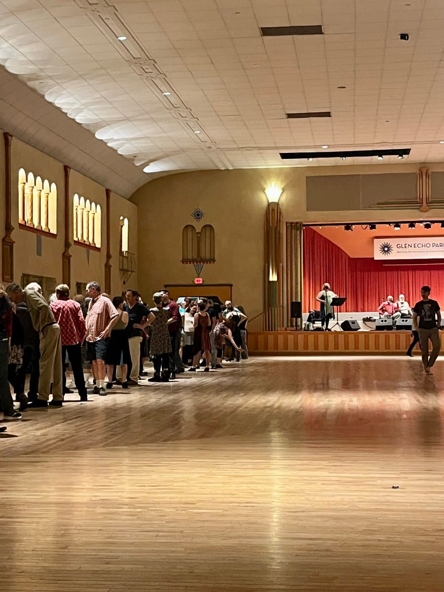 A wide shot of a large indoor hall with a polished wooden floor, where a group of people are standing in a line on the left side. In the background, a stage with a red curtain and a banner is visible, along with a few musicians and other attendees.