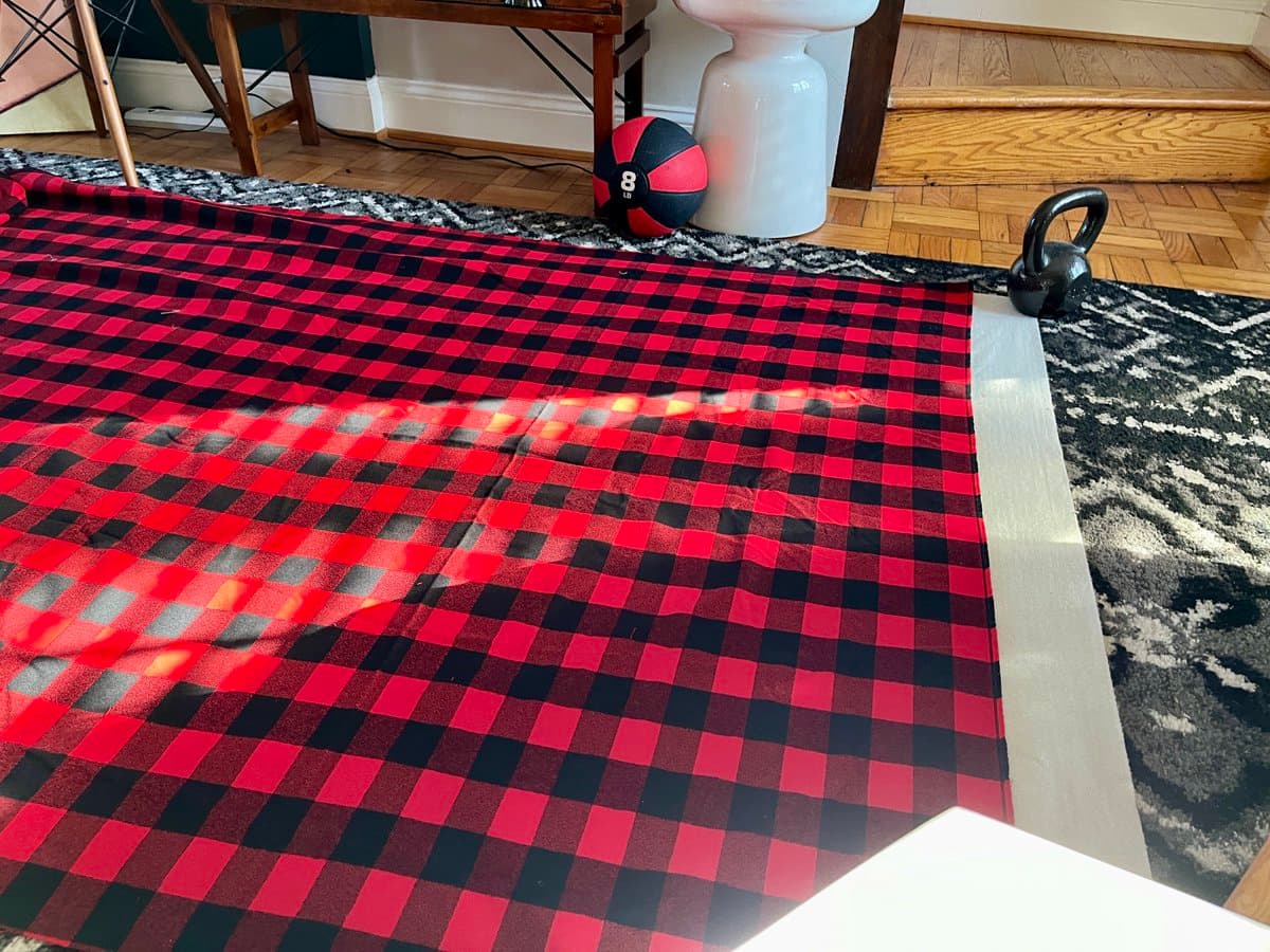 A large piece of red and black buffalo plaid fabric is laid out on a patterned rug on a wooden floor, with a white border visible on the right edge. In the background, a medicine ball and a kettlebell sit near a wooden table and a white ceramic object.