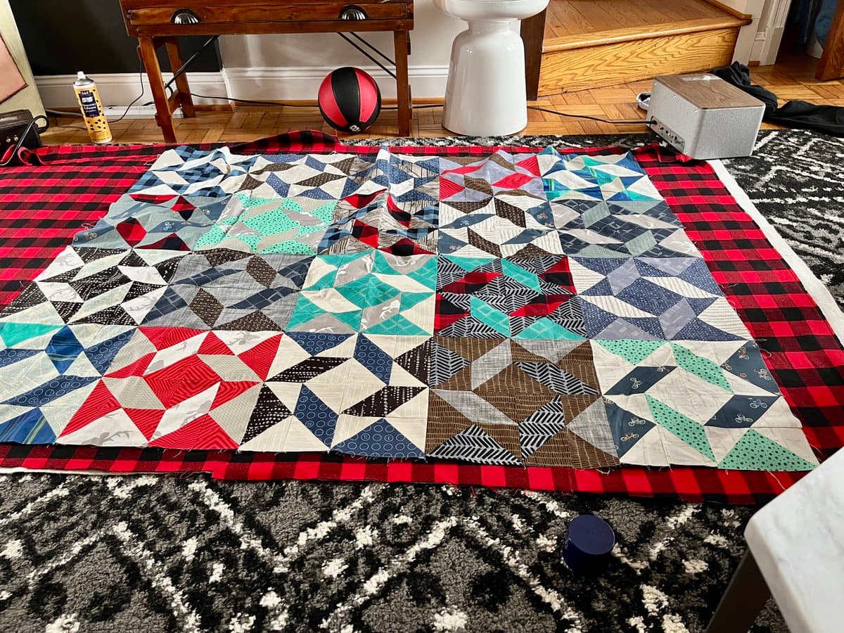 A large, in-progress patchwork quilt with a geometric pattern of triangles and squares in red, blue, green, and brown is laid flat on a red and black plaid backing fabric on a living room floor. The scene includes a wooden table, a red and black ball, a white pedestal, and a gray speaker in the background.