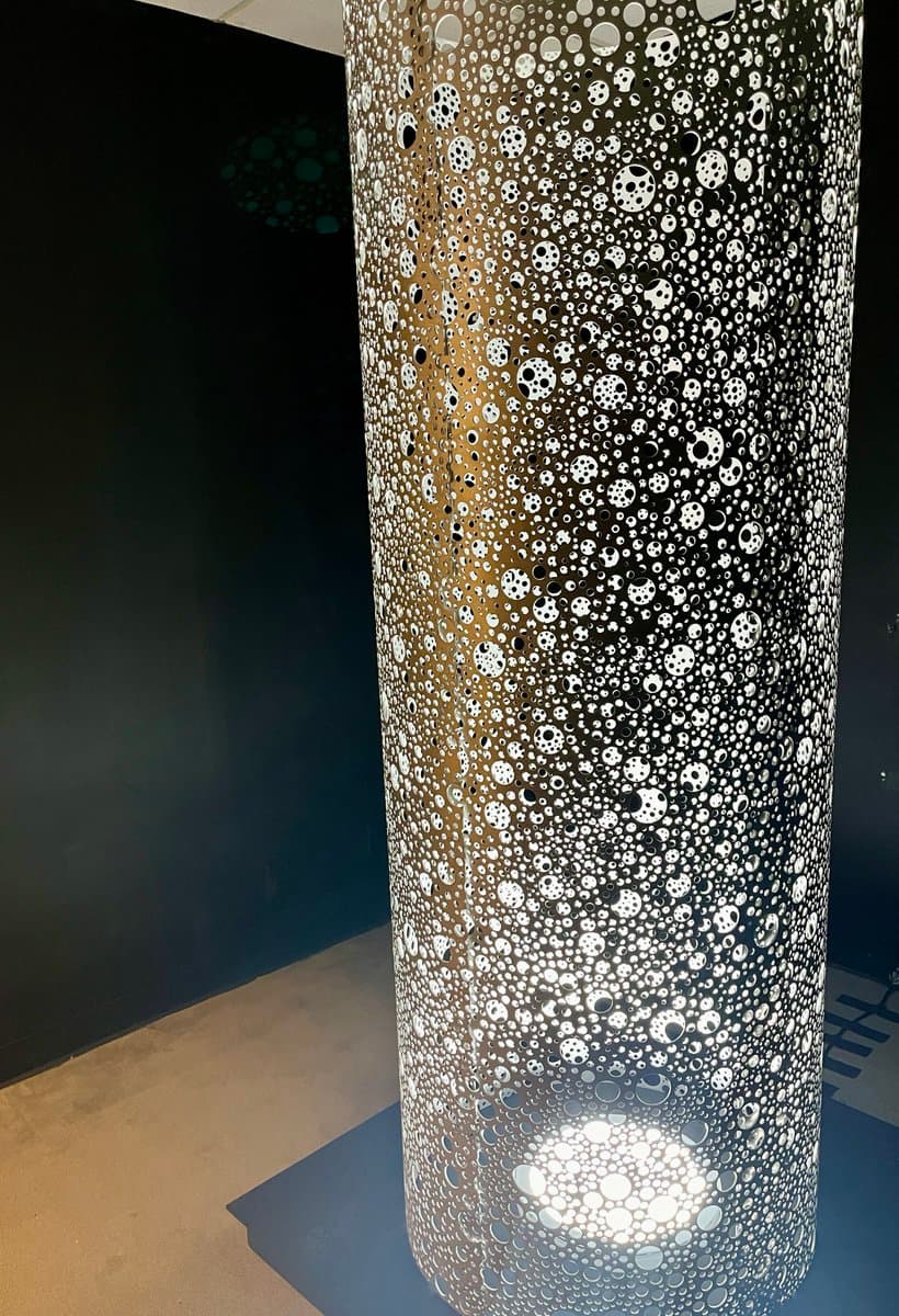 A tall, cylindrical art installation covered in a dense pattern of perforated holes of varying sizes, illuminated from within to create a glowing effect against a dark background.