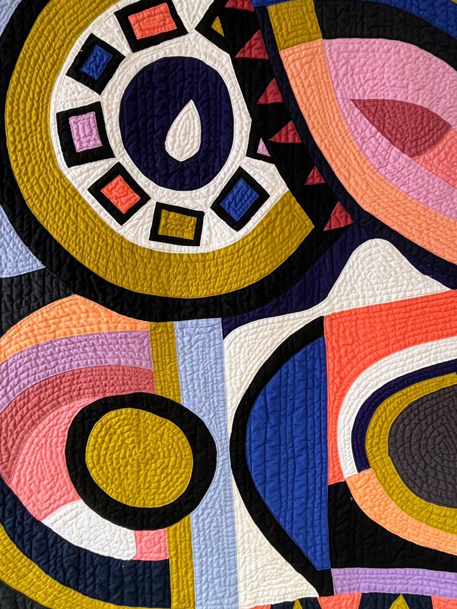 A close-up view of a colorful, abstract quilt featuring bold geometric shapes, including circles, arcs, and squares, in vibrant shades of yellow, pink, blue, and black. The fabric displays a distinct quilted texture with visible stitching lines throughout the design.