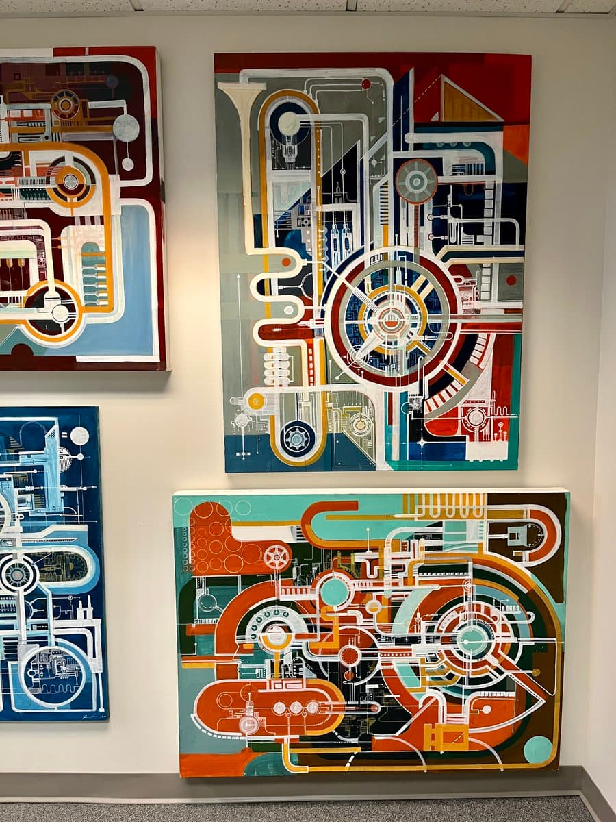 A gallery wall displaying four large, colorful abstract paintings featuring intricate, schematic-like patterns of pipes, gears, and geometric shapes. The artworks are mounted on a plain white wall in an indoor exhibition space.