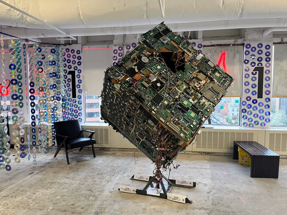 A large, cube-shaped art sculpture made entirely of stacked computer circuit boards and hardware is displayed on a stand in a gallery space. The room features hanging decorations made of CDs and DVDs, a black armchair, and a bench.