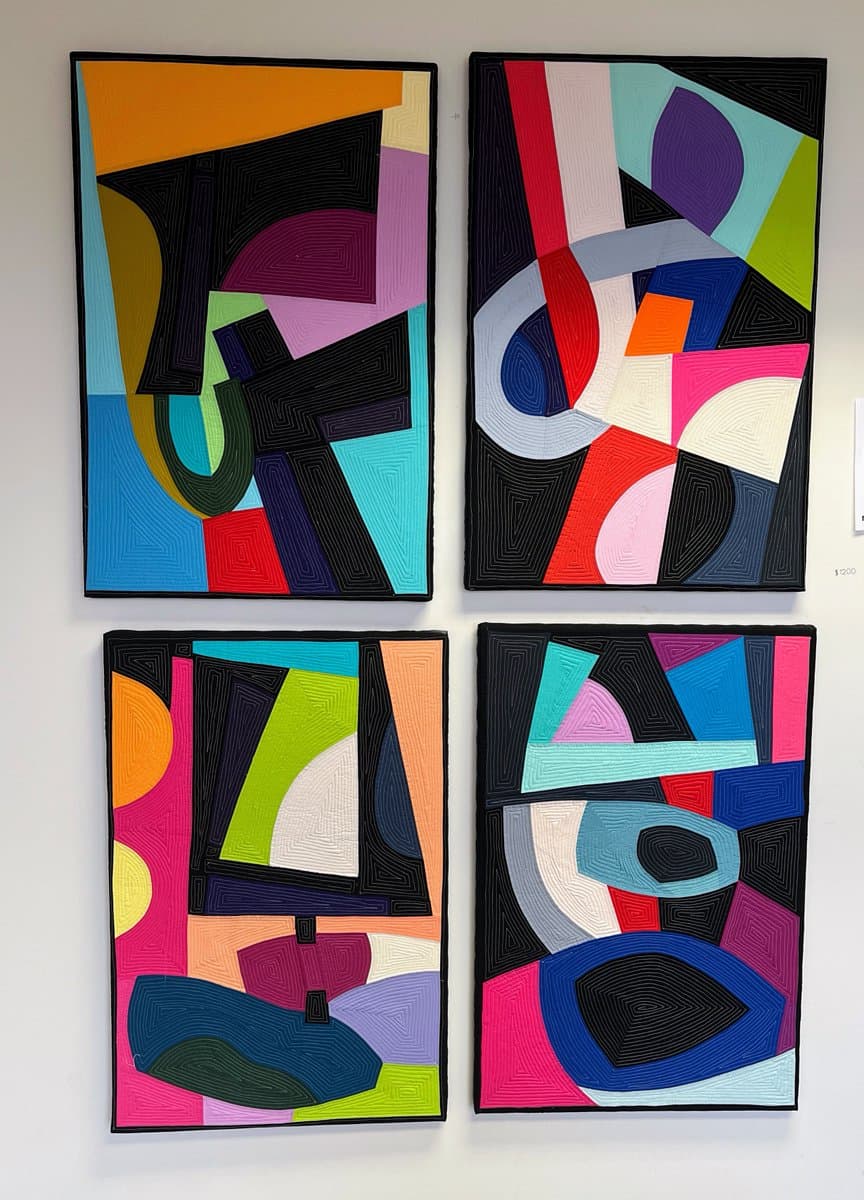 A photograph showing four colorful, abstract paintings arranged in a two-by-two grid on a white wall. The artworks feature bold geometric shapes and thick, textured brushstrokes in a vibrant palette of black, blue, pink, orange, and green.