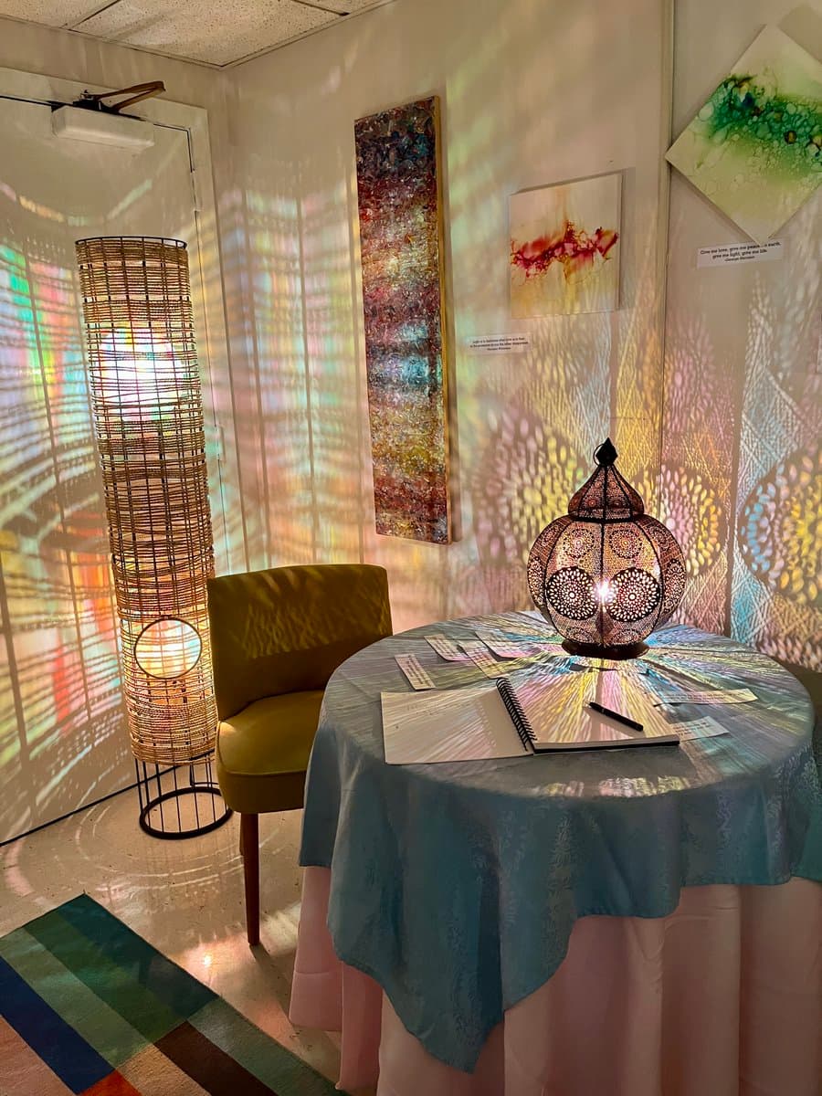 An indoor art installation featuring a round table with a blue tablecloth, a lit Moroccan-style lantern, and a spiral notebook, set against a wall with projected light patterns and hanging artwork. A tall woven floor lamp and a green armchair are visible in the corner. The scene is illuminated by colorful, patterned light projections on the walls and floor.