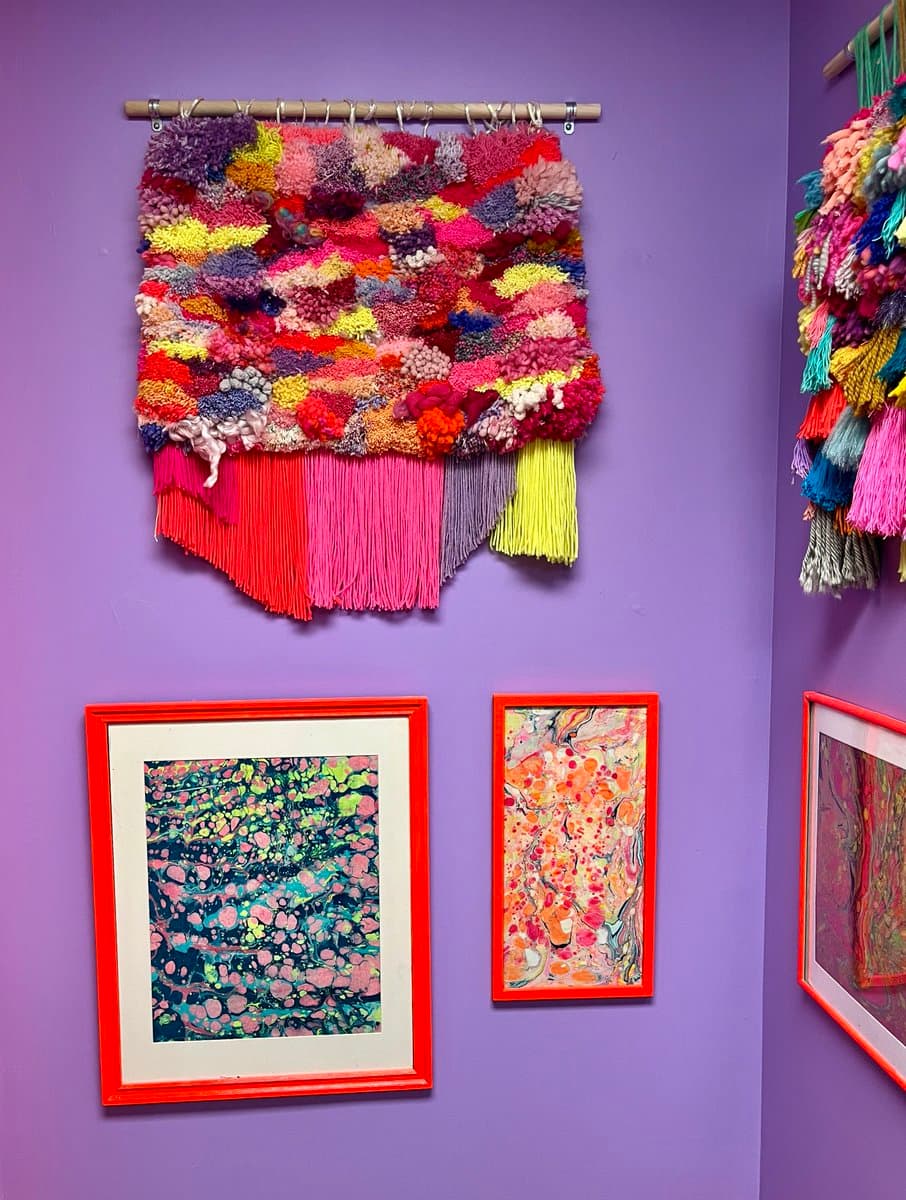 A gallery wall painted purple, displaying a colorful, textured fiber art piece with fringe hanging from a rod, alongside two framed abstract artworks with orange frames.