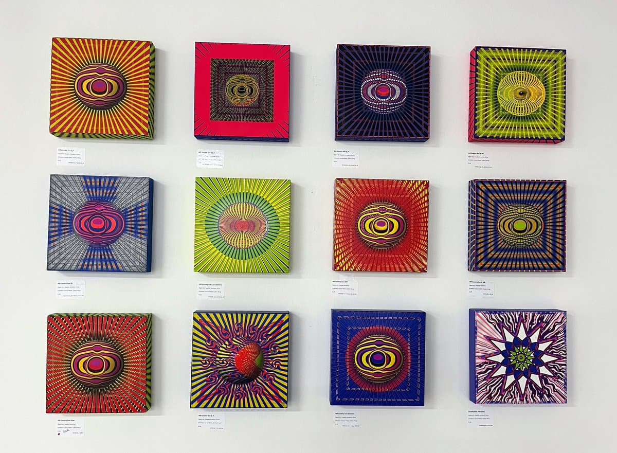 A gallery wall displaying twelve square, three-dimensional artworks arranged in a 3x4 grid. Each piece features vibrant, geometric, and psychedelic patterns with radiating lines and concentric shapes in various colors. Small, illegible white labels are placed on the wall beneath each artwork.
