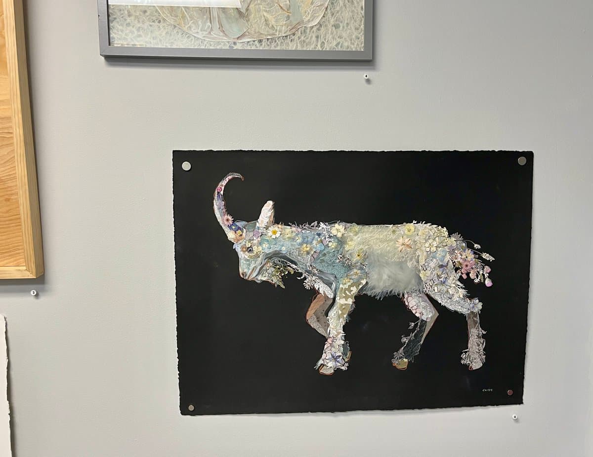 A photograph of a mixed-media art piece displayed on a gallery wall, featuring a goat-like animal constructed from small flowers and textured materials against a black background. The artwork is mounted with silver pins, and portions of two other framed pieces are visible at the top and left edges of the frame.