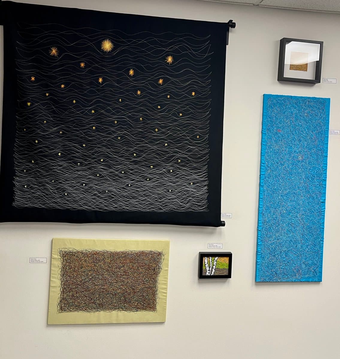 A gallery wall displaying several textile art pieces, including a large black hanging with white wavy lines and gold star-like accents, a tall blue textured piece, a smaller brown and black abstract piece, and two small framed artworks.