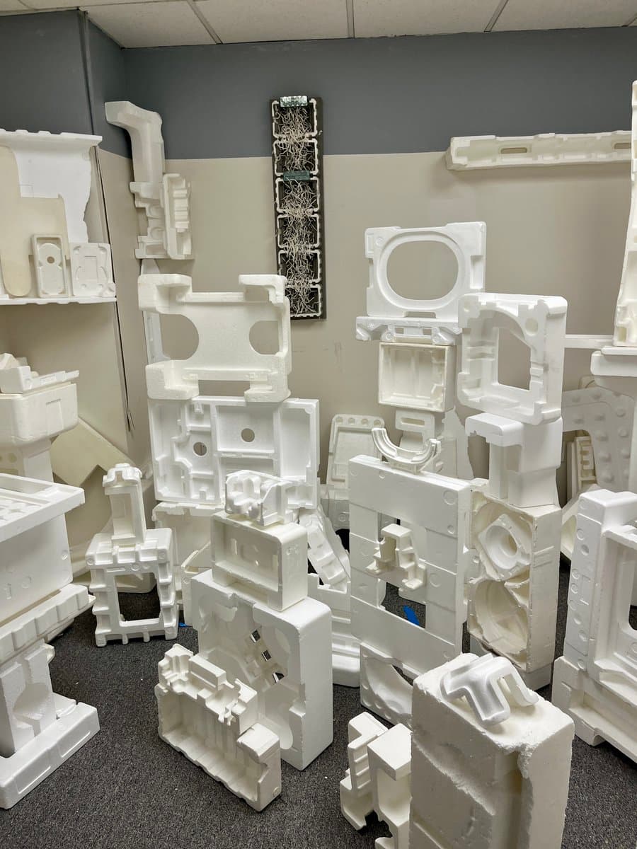 A room filled with numerous white, molded plastic or foam components stacked on the floor and shelves, likely parts for appliances or machinery. The pieces have various shapes with cutouts, ridges, and mounting points, suggesting they are in a manufacturing or assembly area.