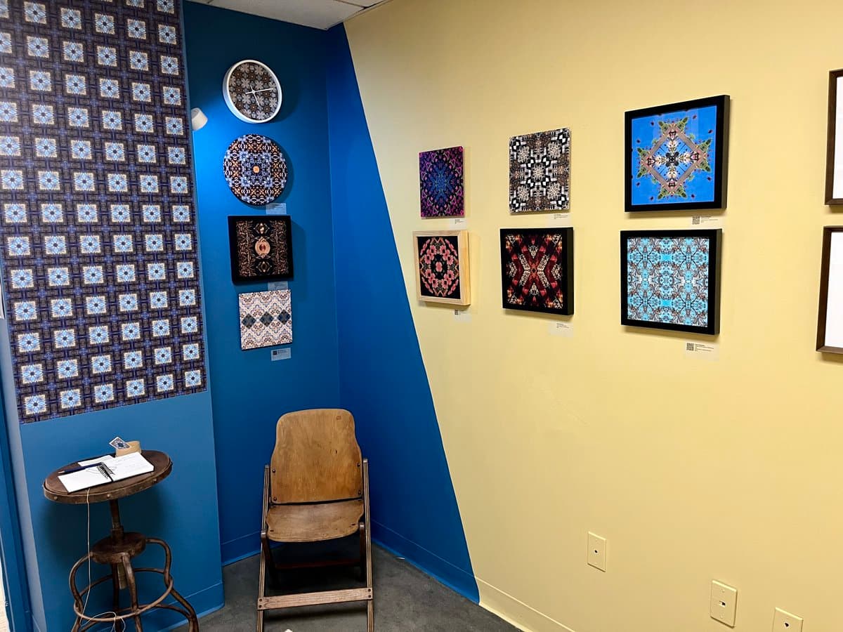 An art gallery corner featuring a blue wall and a yellow wall, both displaying various framed and unframed pieces of geometric and patterned artwork. A wooden chair and a small round table with a notebook sit on the floor in the foreground.