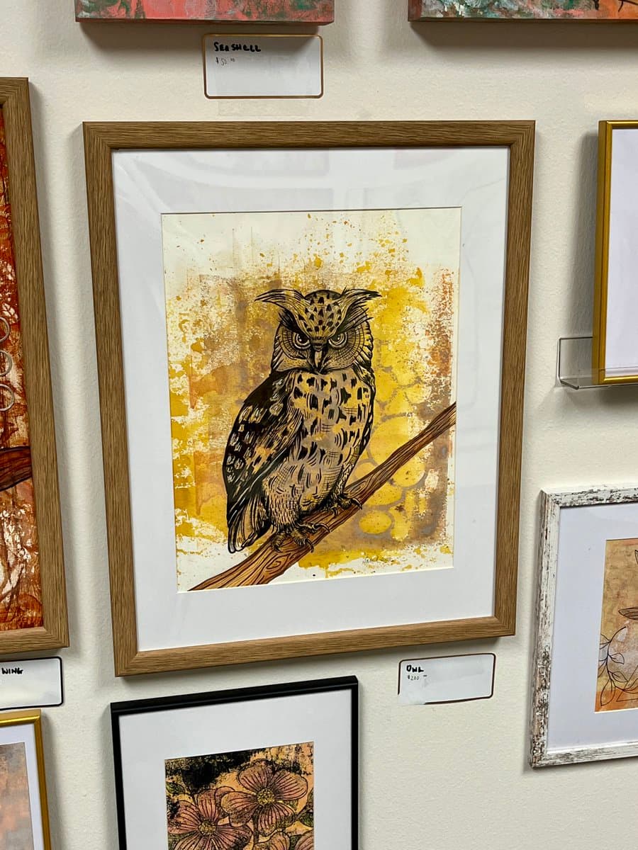 A framed art print of an owl perched on a branch, displayed on a gallery wall among other framed artworks. The owl is depicted in a detailed, illustrative style against a textured yellow and white background. Small price tags are visible on the wall, including one labeled 'OWL $220' below the main piece.