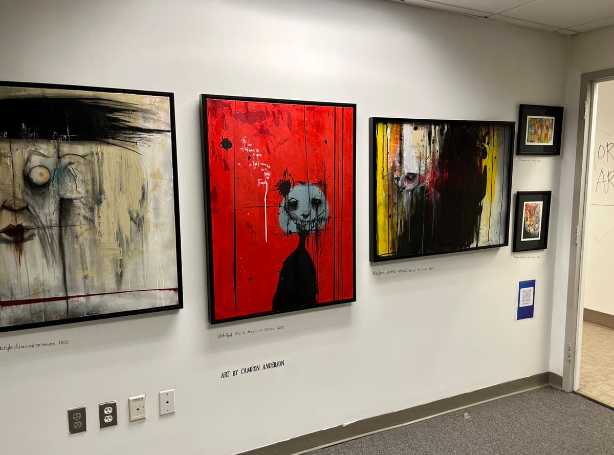 A gallery wall displaying three large, framed abstract paintings with expressive, dark imagery, along with two smaller framed pieces on the right. Below the central red painting, a label reads "ART BY CAMRON ANDERSON."