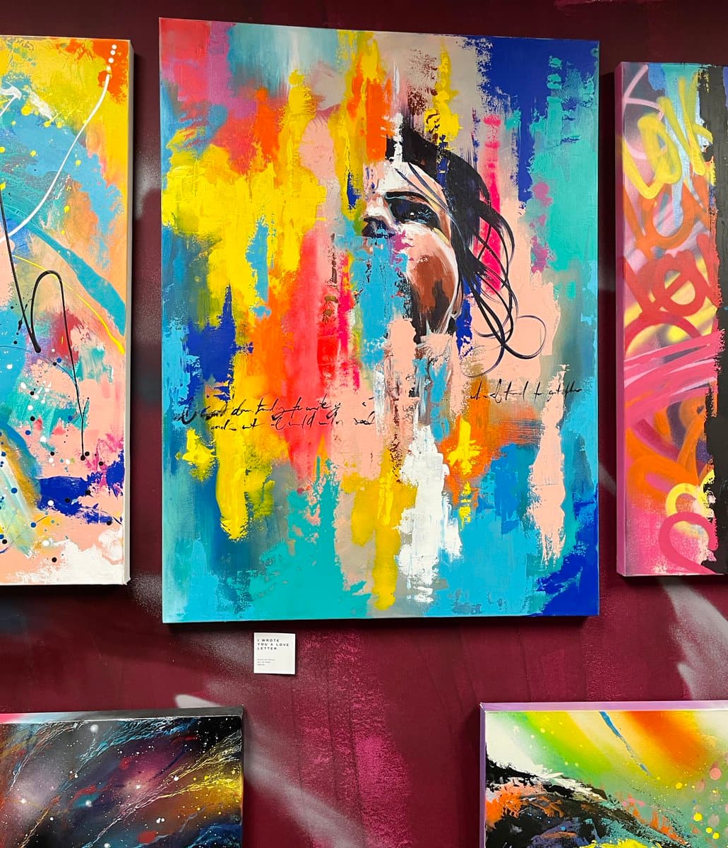 A photograph of several colorful abstract paintings displayed on a dark red wall, with a central piece featuring a stylized portrait of a woman. A small white label is visible on the wall below the central artwork.