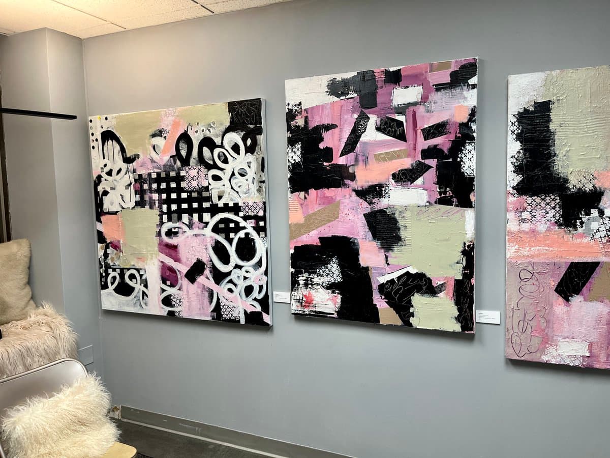 A photograph of three abstract paintings hanging on a gray wall in an indoor setting, featuring bold brushstrokes in black, white, pink, and beige. A white furry chair is partially visible on the left side of the frame.