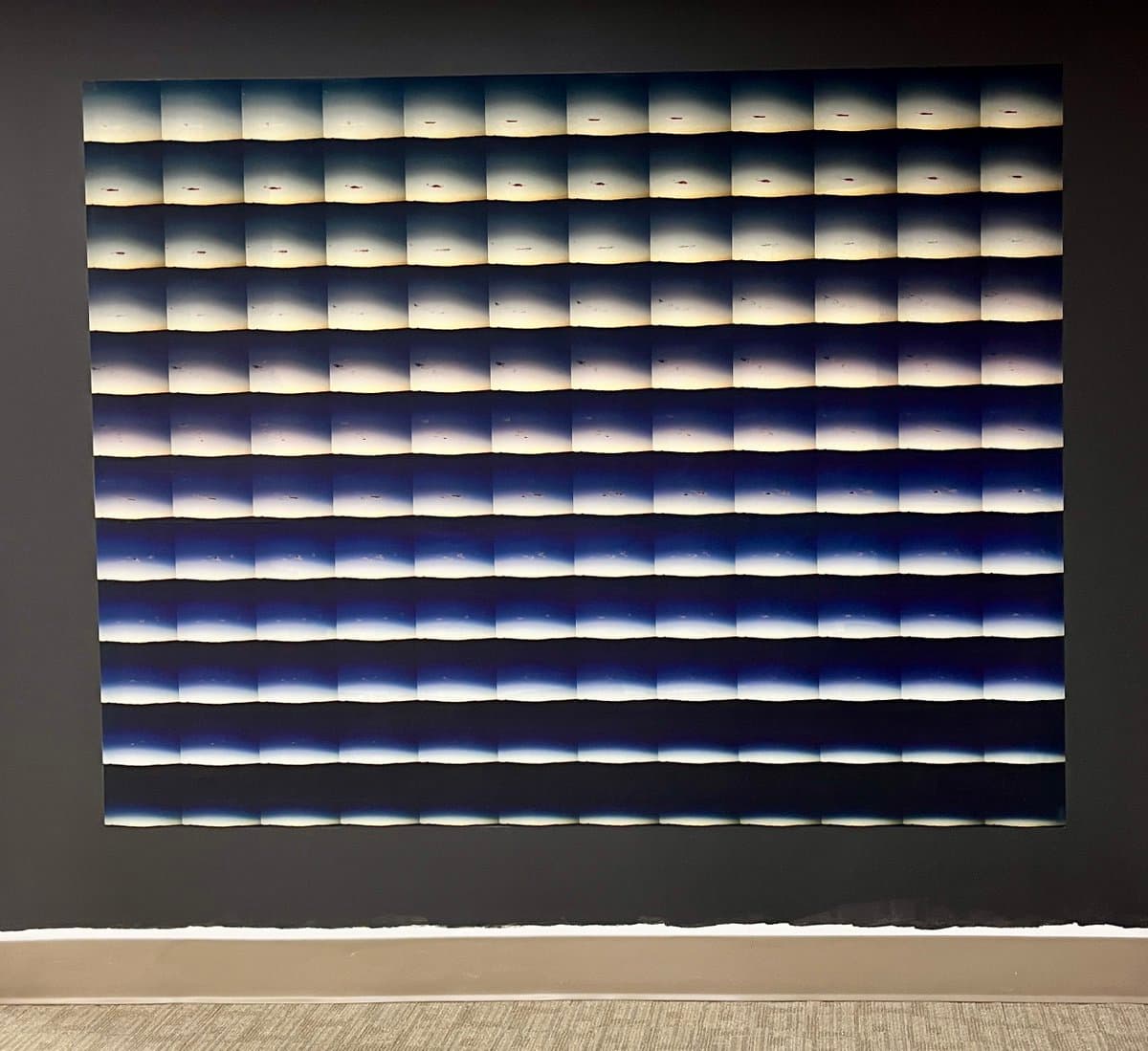 A large, square art installation mounted on a dark wall, featuring a grid of repeating rectangular panels that display a gradient from dark blue to white, resembling a stylized horizon or sky. The piece is displayed in a gallery setting with a light-colored floor and baseboard visible at the bottom.