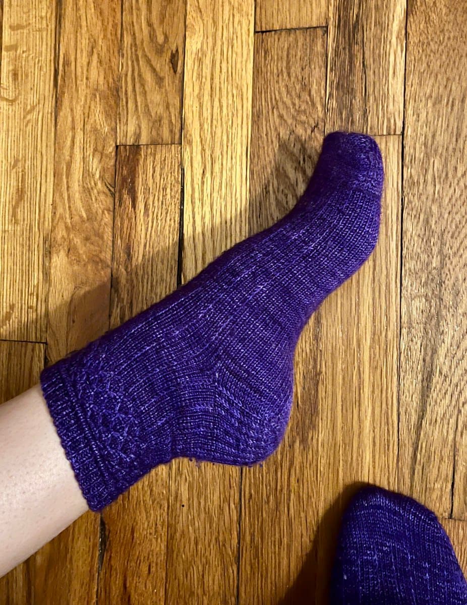 A top-down photograph of a person's foot wearing a purple knitted sock, resting on a wooden floor. The texture of the knit and the wood grain are clearly visible.