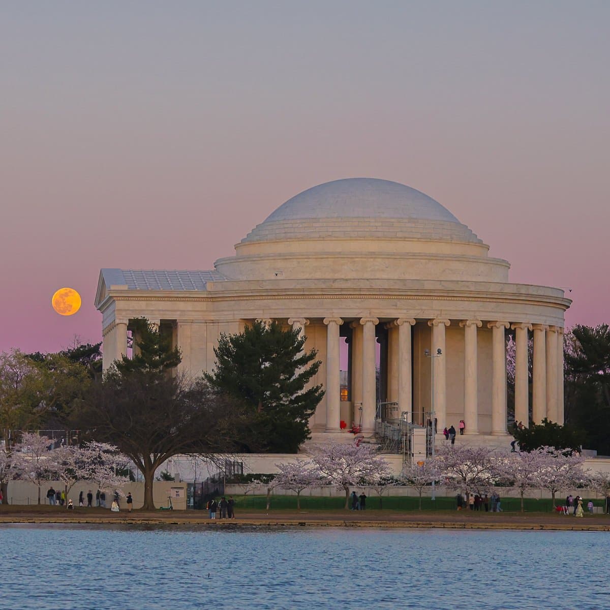 A scenic photograph of the Jefferson Memorial in Washington, D.C., taken at dusk or dawn. A large, bright orange full moon is visible in the sky to the left of the building, with cherry blossom trees in the foreground along the waterfront.