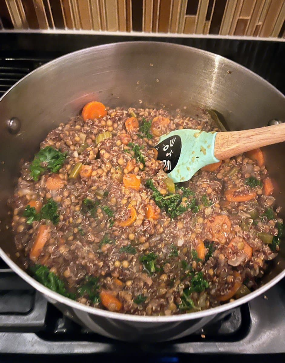 A close-up, top-down view of a stainless steel pot on a stovetop containing a simmering lentil and quinoa stew with visible chunks of carrot and green leafy vegetables. A wooden-handled spatula with a light blue silicone head rests in the pot.