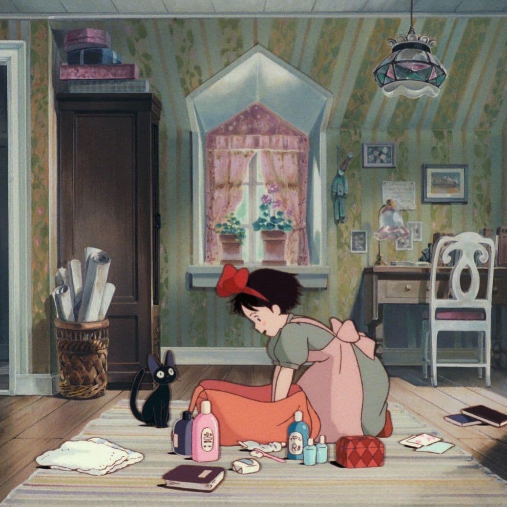 A still from the 1989 animated film Kiki's Delivery Service, showing a young girl with a red bow sitting on a rug in a room with a black cat and various items on the floor.