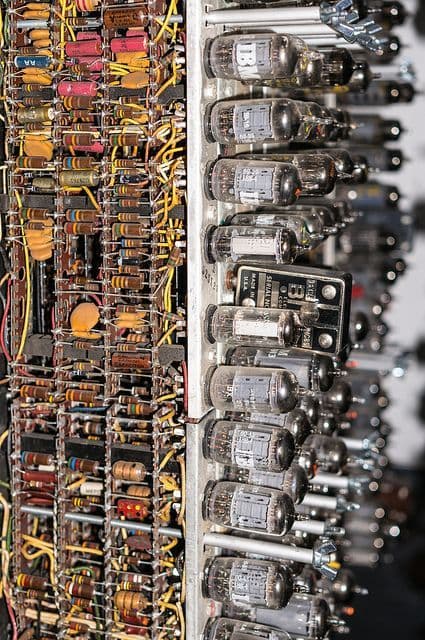 A close-up photograph of a vintage electronic circuit board, featuring rows of glass vacuum tubes on the right and a dense array of resistors, capacitors, and wiring on the left.
