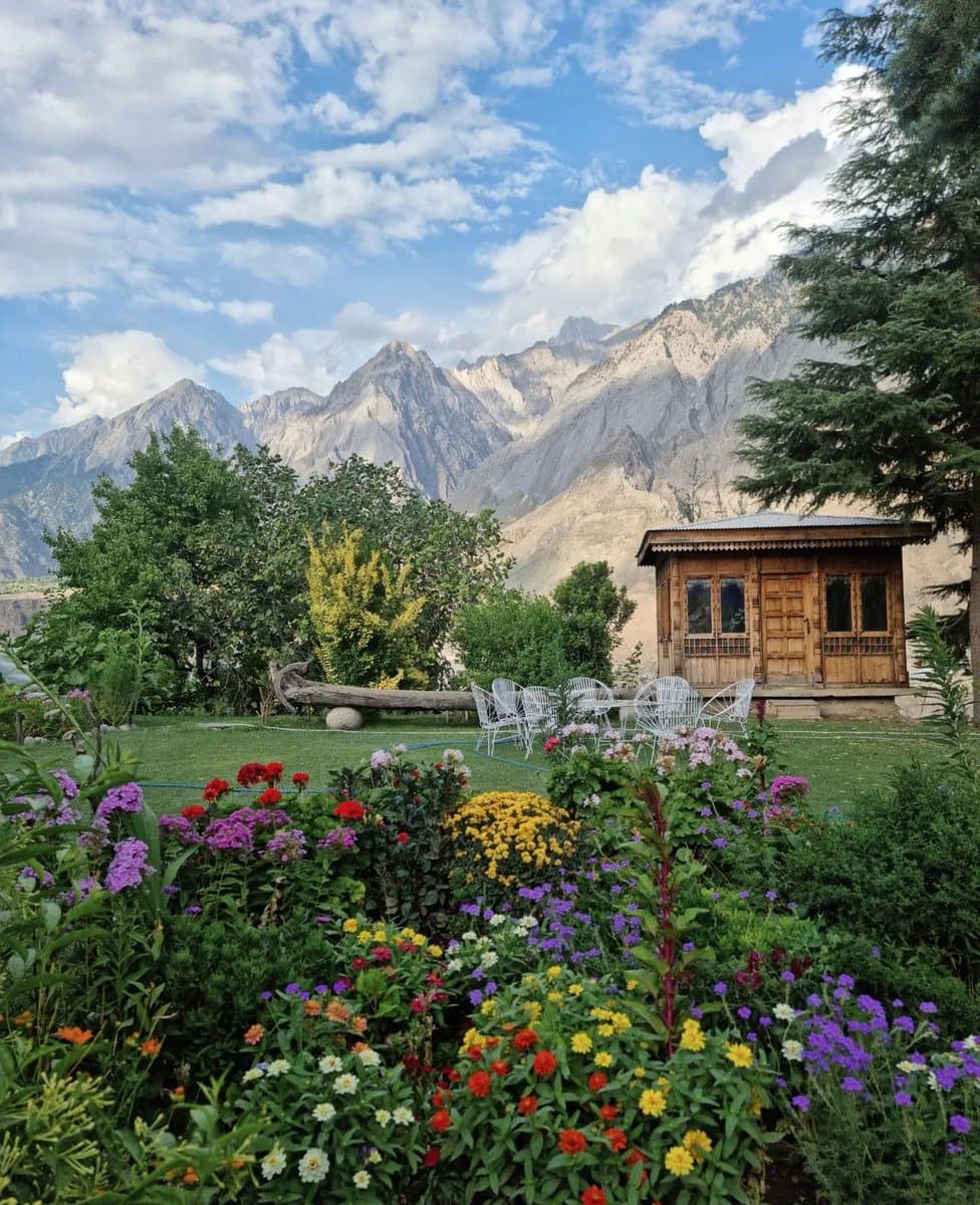 A scenic landscape photograph showing a vibrant flower garden in the foreground, with a wooden cabin and white chairs on a lawn in the midground, and a rugged mountain range under a partly cloudy sky in the background.