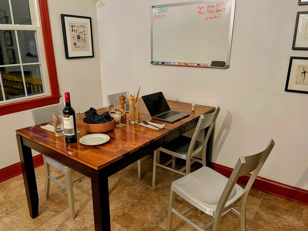 A photo of a dining room featuring a wooden table set with a bottle of red wine, glasses, and a laptop. A whiteboard on the wall displays a handwritten weekly schedule.