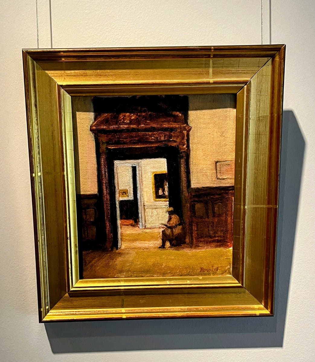 A framed oil painting hanging on a white gallery wall, depicting an interior scene with a seated figure in a doorway. The artwork is enclosed in a thick, ornate gold frame.