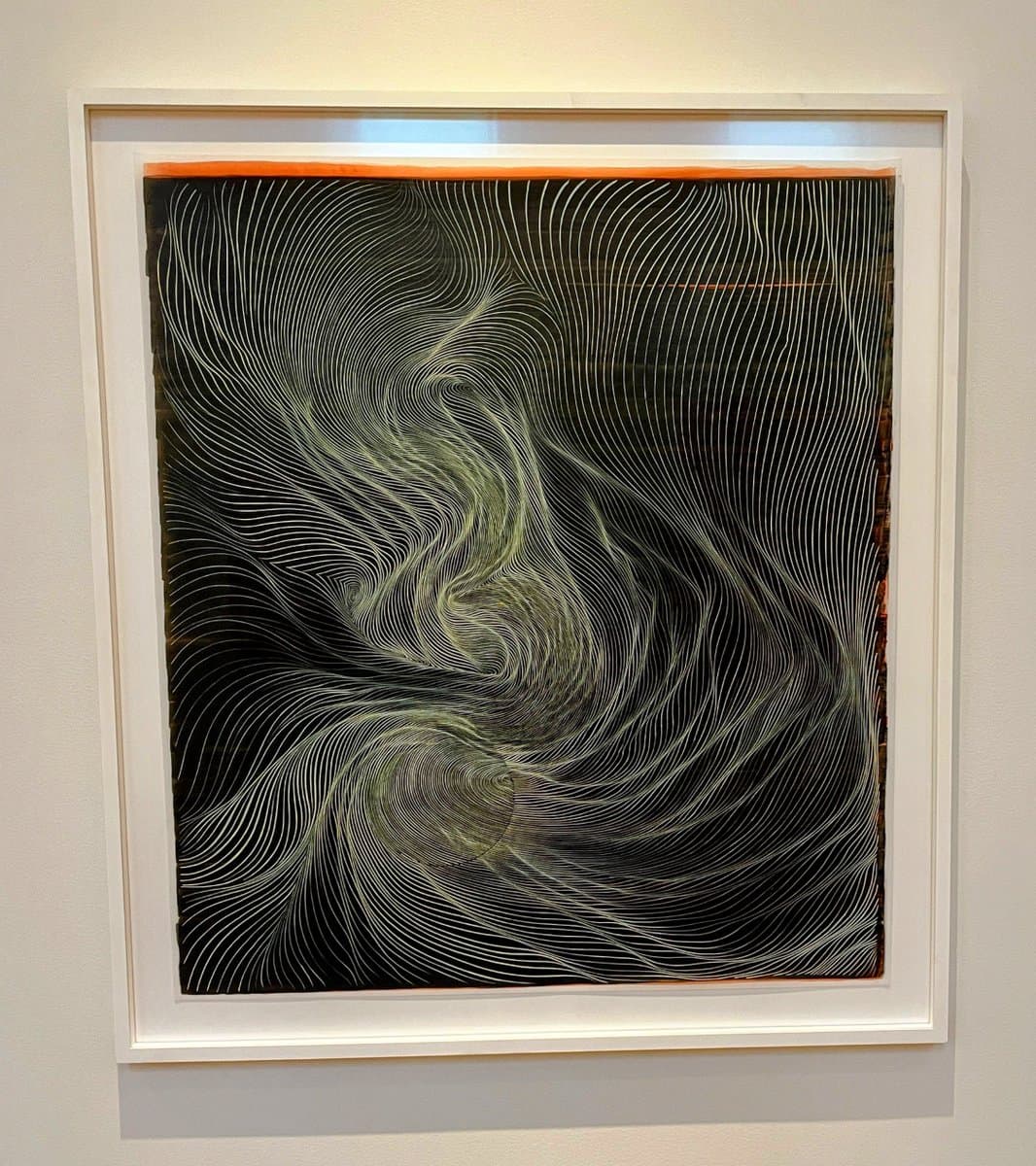 A framed abstract artwork hanging on a white gallery wall, featuring a black background with intricate, swirling white and pale green lines that create a sense of depth and movement. The piece is set within a white frame with a thin orange border at the top.