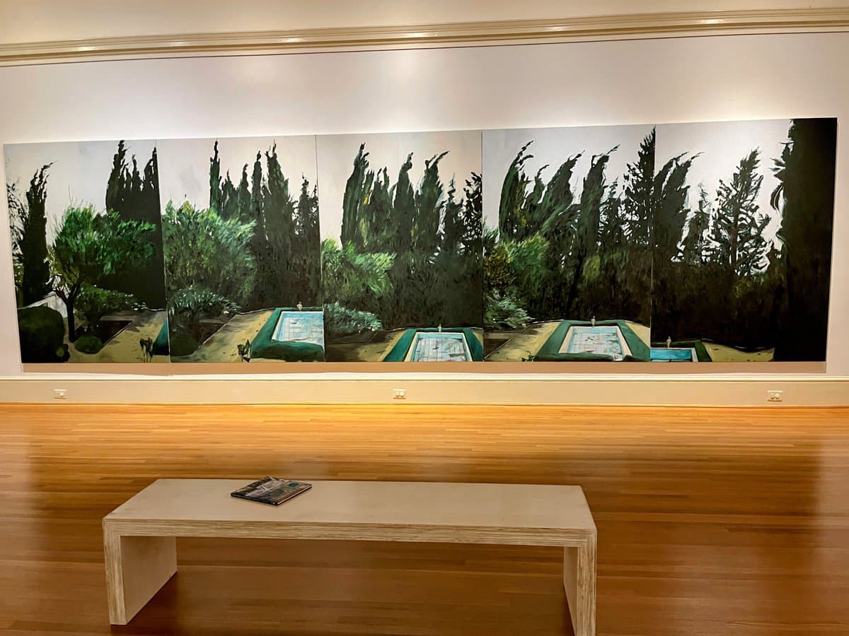 A wide, multi-panel painting depicting a landscape with tall, dark trees and a swimming pool is displayed on a white gallery wall. In the foreground, a low wooden bench sits on a polished wood floor, with a small booklet resting on its surface.