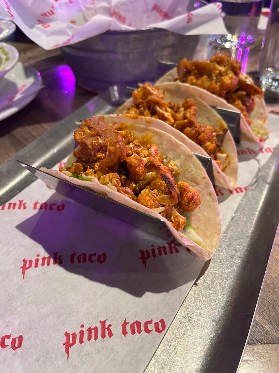 A close-up photograph of three tacos filled with seasoned meat and vegetables, served in metal taco holders on a tray lined with branded paper. The paper features the text "pink taco" printed repeatedly in red. The setting appears to be a restaurant with purple ambient lighting.