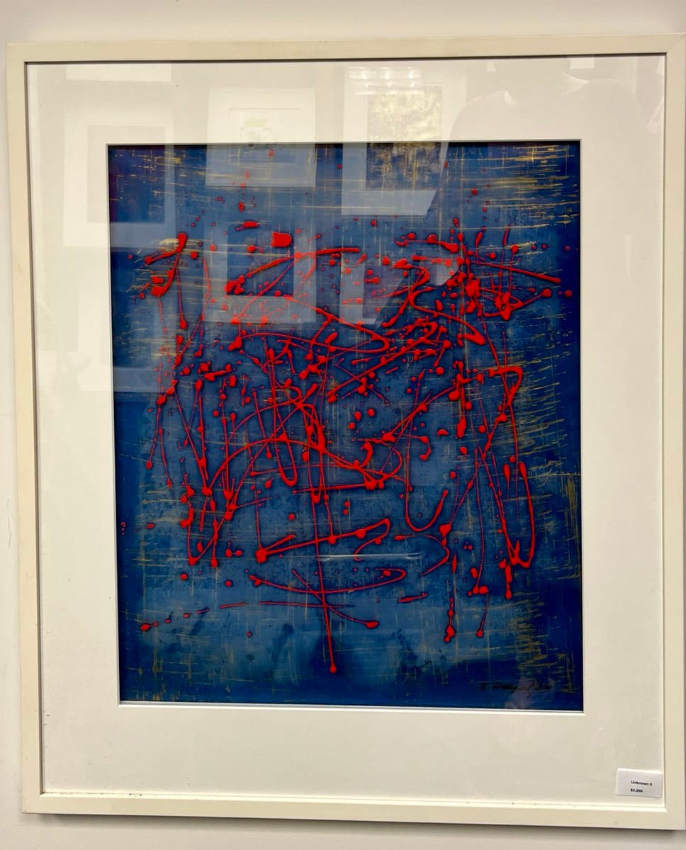 A framed abstract painting featuring a deep blue background with chaotic, splattered red lines and dots. The artwork is displayed behind glass, which shows reflections of the surrounding room, and includes a small price tag in the bottom right corner.