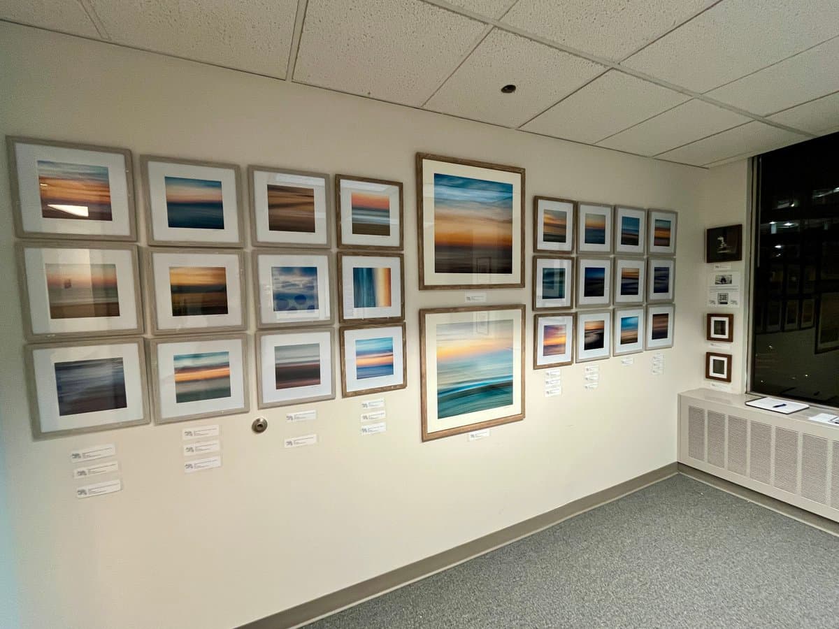 A gallery wall displaying a collection of framed landscape photographs, primarily featuring blurred, long-exposure shots of sunsets and sunrises over water. The images are arranged in a grid pattern with small informational placards beneath each frame.
