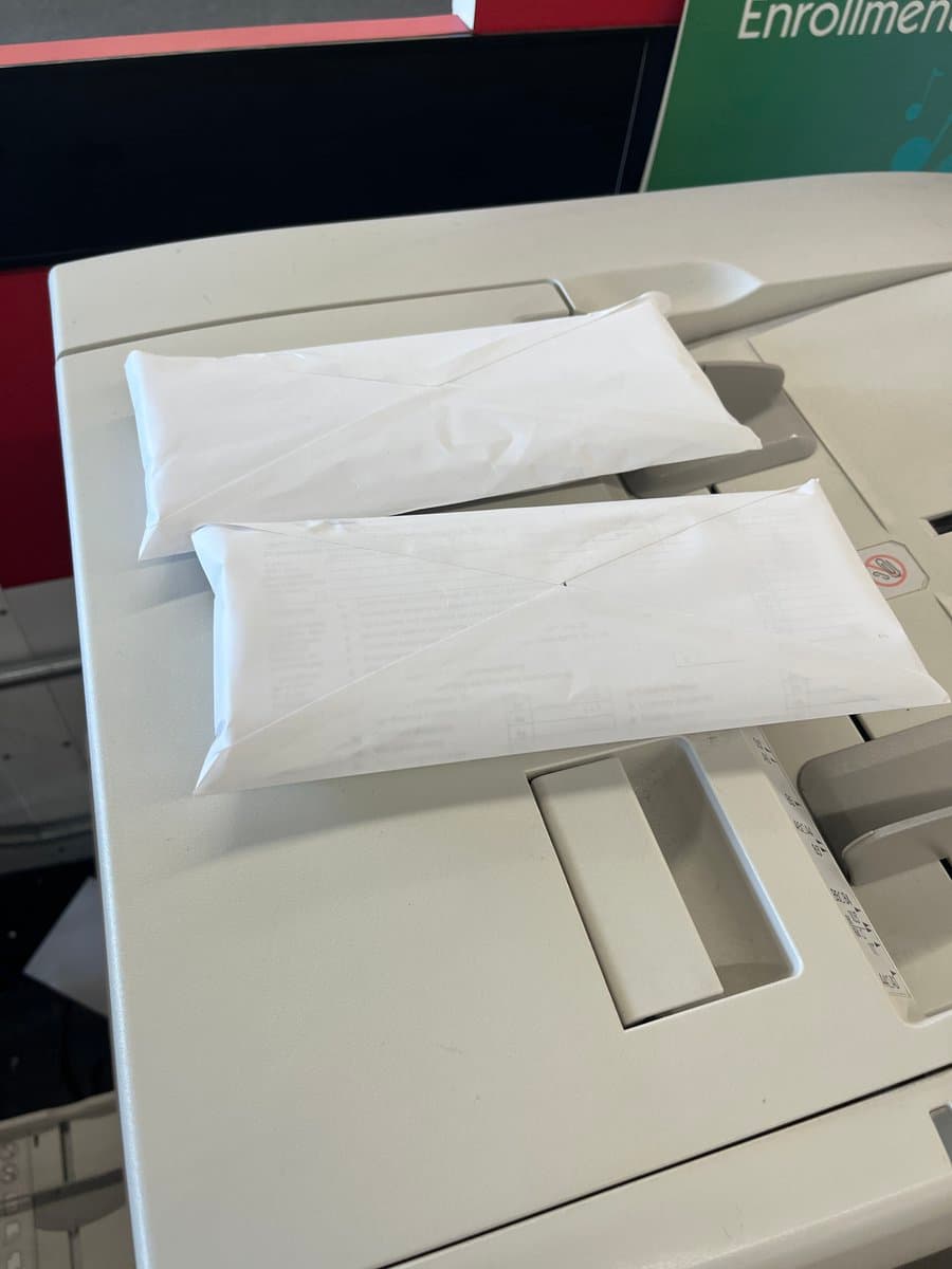 Two thick, white paper envelopes containing printed documents sit on the output tray of an office printer. In the background, a green sign is partially visible with the word 'Enrollment' printed on it.