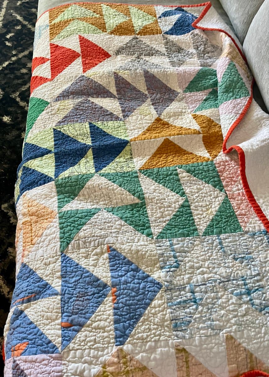 A close-up photograph of a handmade patchwork quilt featuring a geometric pattern of triangles in various colors, including red, blue, green, and yellow, draped over a piece of furniture.