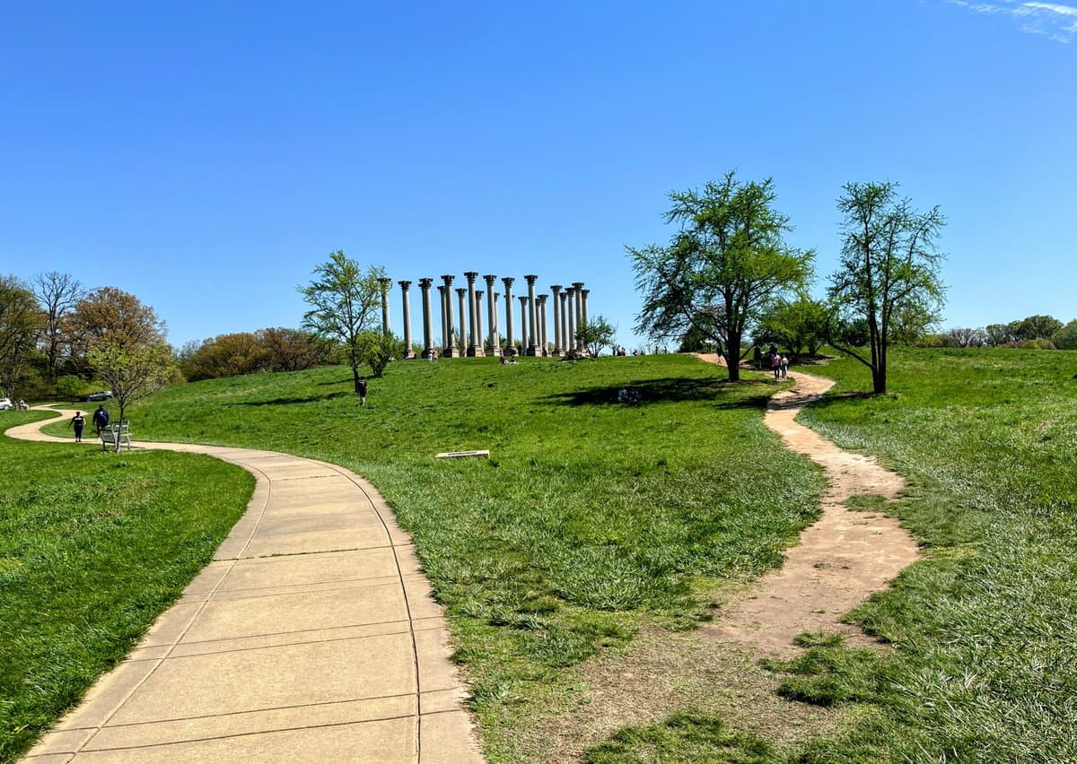 A scenic outdoor photograph of a park on a sunny day, featuring a paved walking path and a dirt trail winding through a grassy hill toward a monument made of tall stone columns. Several people are visible walking along the paths and near the monument, with trees scattered across the landscape under a clear blue sky.