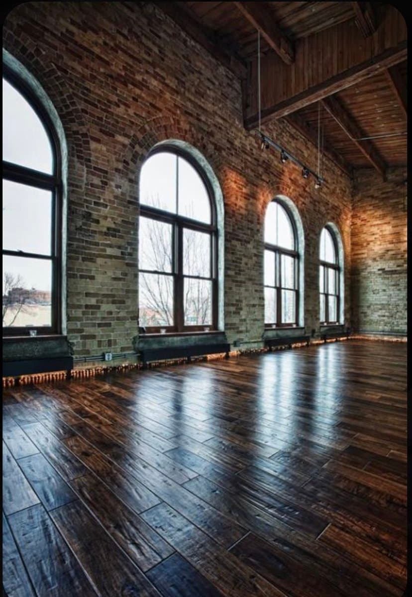 A photograph of a spacious, empty interior room with exposed brick walls, a polished dark wood floor, and a high wooden-beamed ceiling. A row of large arched windows lines one wall, letting in natural light that reflects off the floor.