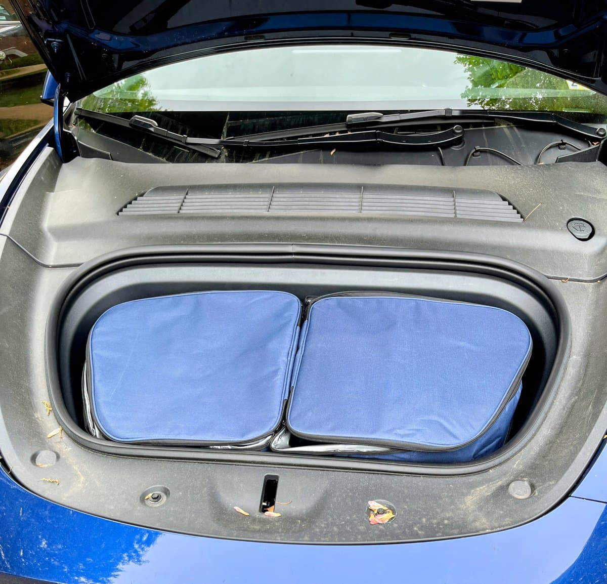 A photo of the open front trunk (frunk) of a blue car, containing two blue, zippered cooler bags placed side-by-side. The car's windshield and wipers are visible in the background.