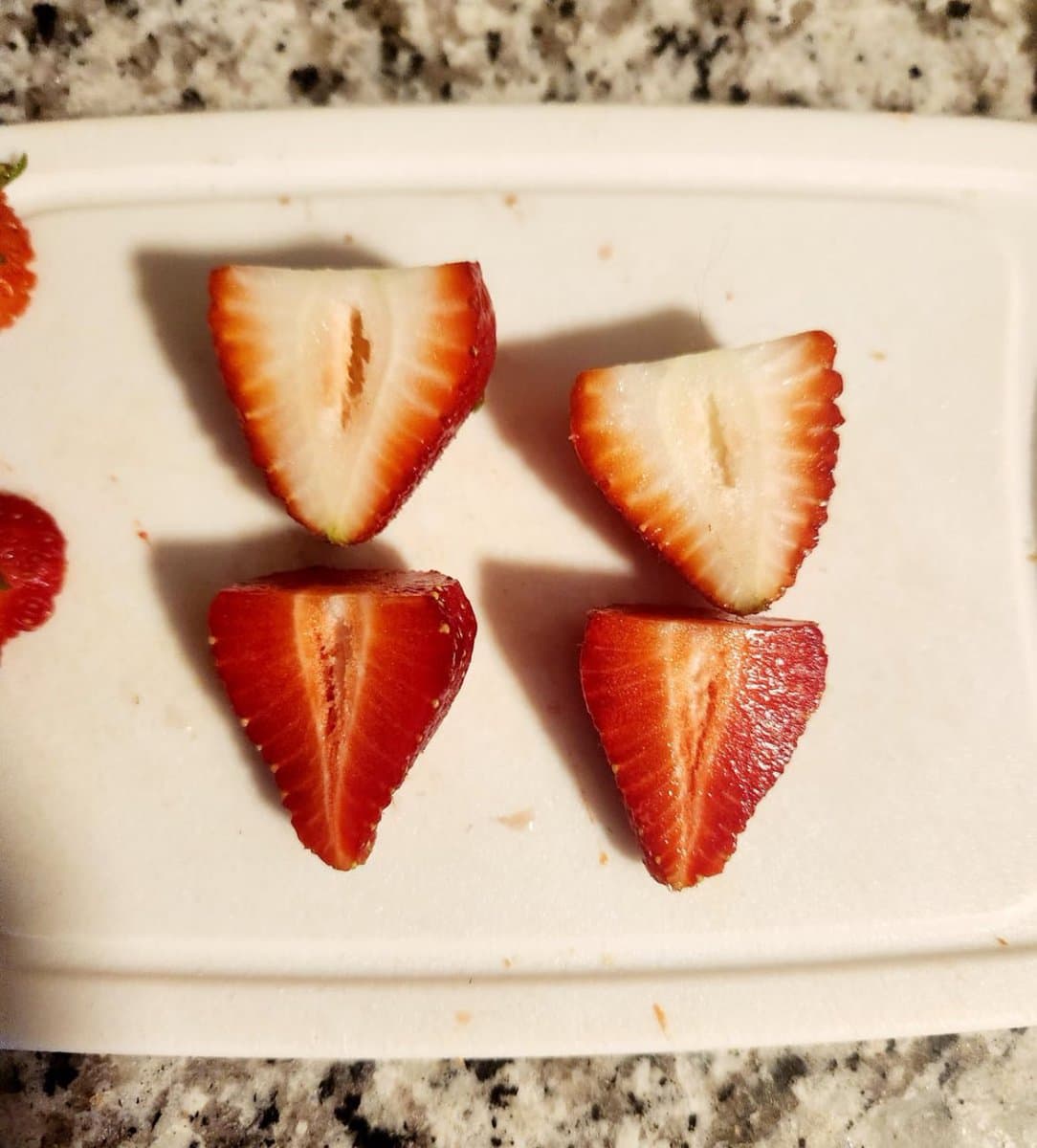 A close-up photograph of four strawberry halves arranged on a white cutting board, showing a contrast between pale, white-fleshed berries and deep red ones. The board rests on a speckled granite countertop.