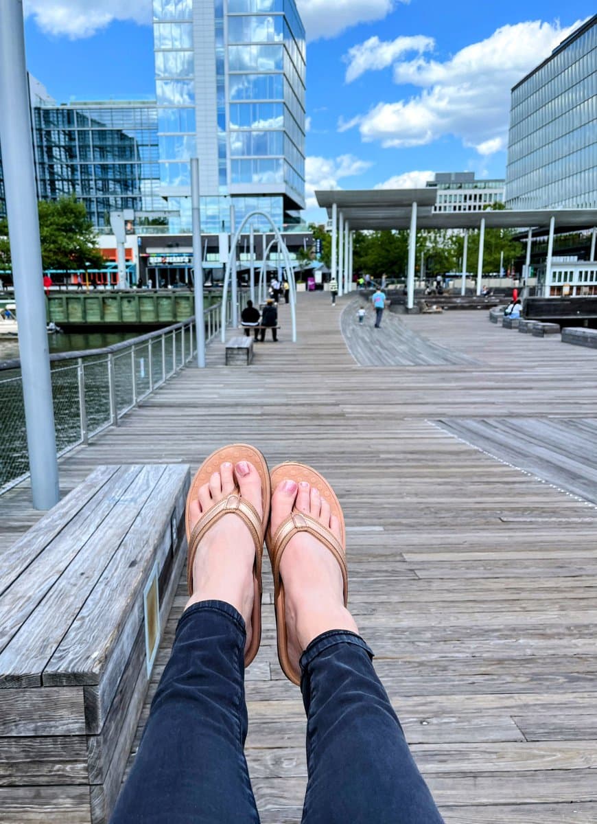 A first-person perspective photo of a person's feet in tan flip-flops resting on a wooden bench at a waterfront promenade, with modern glass buildings and a blue sky in the background.