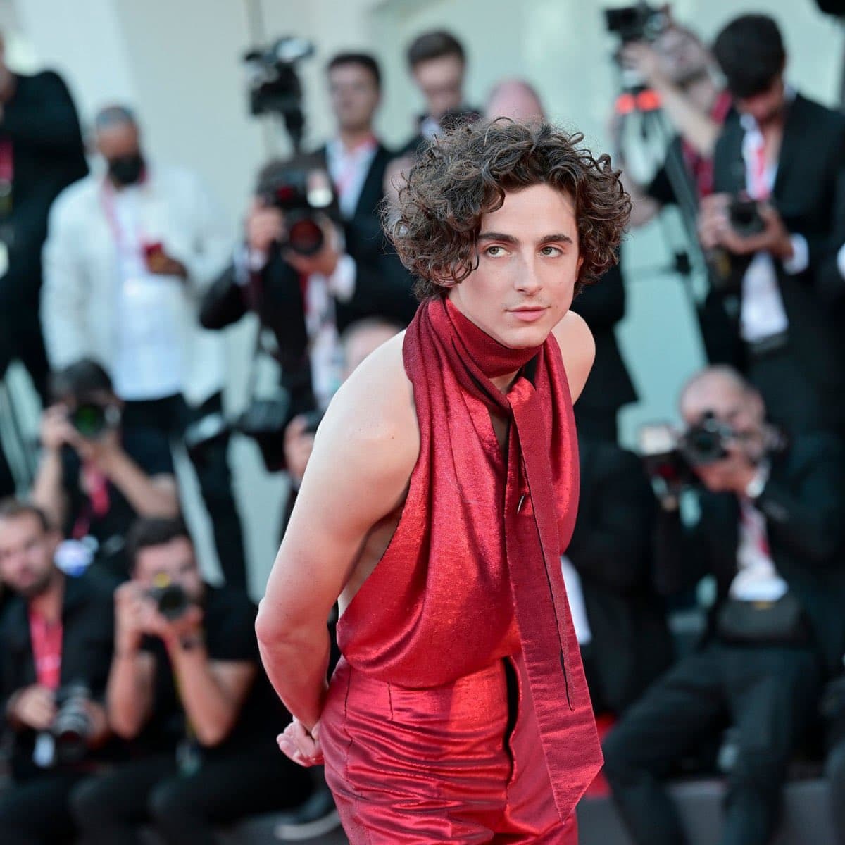 A photograph of a young man with curly brown hair wearing a sleeveless, metallic red outfit and a matching scarf, posing on a red carpet. In the blurred background, numerous photographers and event staff are visible, indicating a high-profile public event.