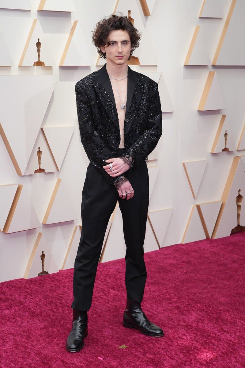 A full-length photograph of a young man with curly dark hair standing on a red carpet in front of a white backdrop featuring Oscar statuette logos. He is wearing a black sequined blazer over a bare chest, black trousers, and black leather boots.