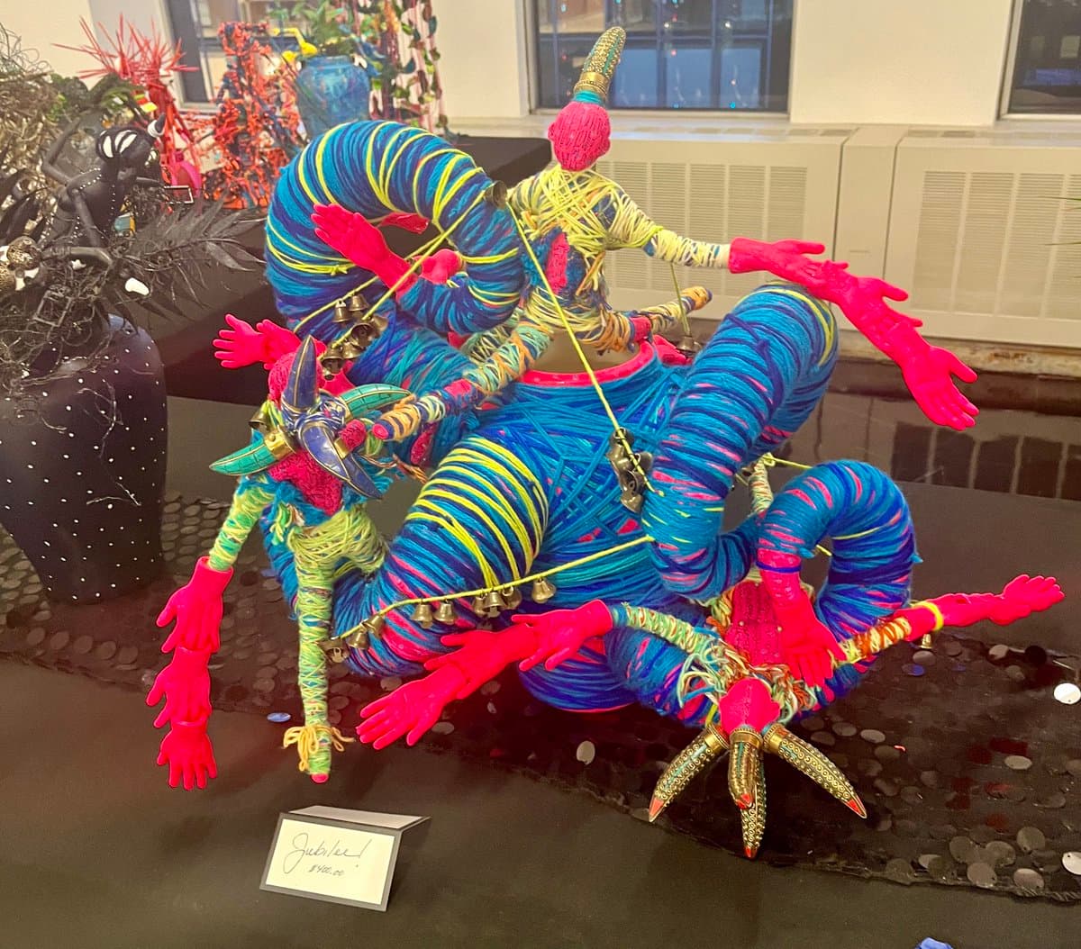 A vibrant, multi-armed sculpture made of wrapped yarn in bright blue, pink, and yellow, adorned with bells and metallic accents, displayed on a table. A small handwritten price tag in the foreground reads 'Jubilee! $40.00'.